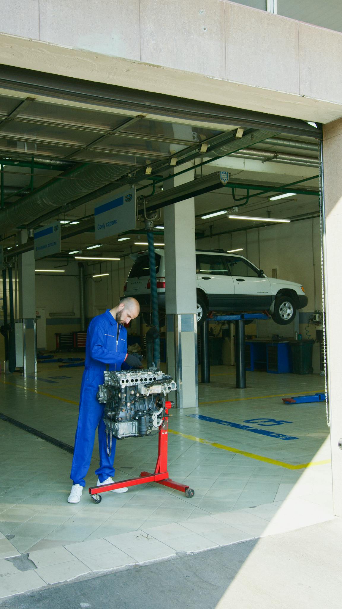 Mechanic in blue uniform inspecting an engine in a bright auto repair shop. Car lifted in background.